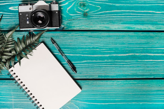 Green fern leaves; spiral notepad; pen and camera against wooden turquoise background