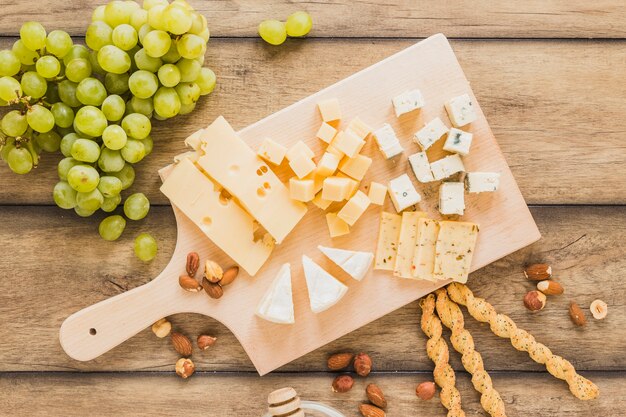 木製の机の上のまな板の上の緑のブドウ、アーモンド、パンの棒およびチーズブロック