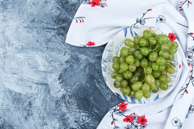 Uva verde in un piatto sul fondo grungy dell'intonaco e dell'asciugamano da cucina. laici piatta.