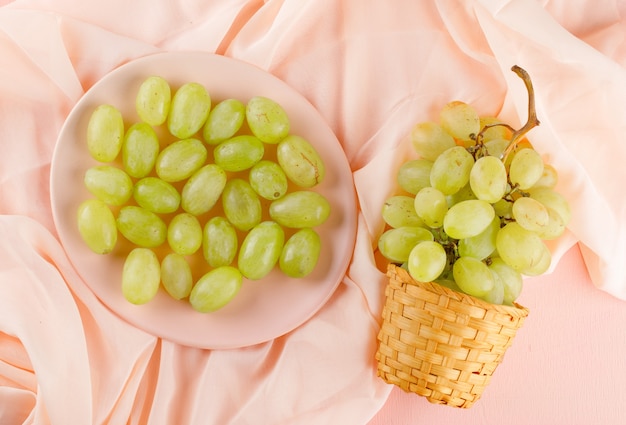 Green grapes in wicker basket and plate flat lay on a pink textile