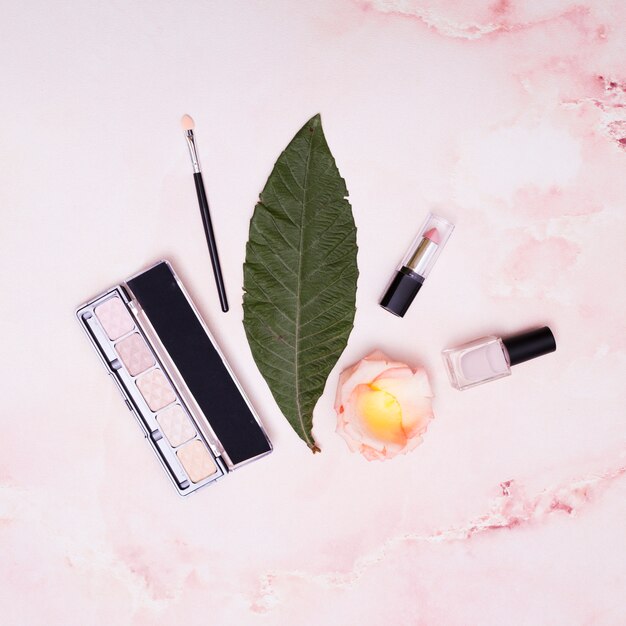 Green leaf; lipstick; nail varnish; petals; makeup brush and eyeshadow palette on pink backdrop