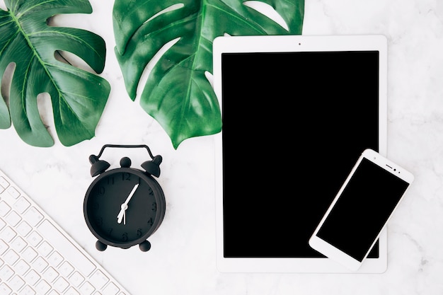 Free photo green monstera leaves; alarm clock; keyboard; digital tablet and mobile phone on textured background