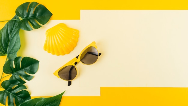 Green monstera leaves with sunglasses and scallop on paper against yellow background