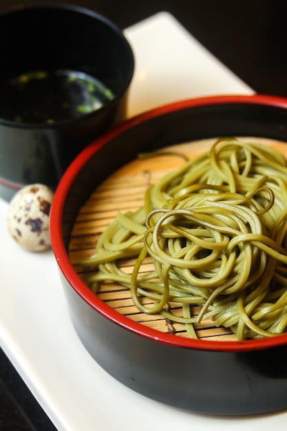 Green noodles on a black bowl with quail egg and souse on a white tray