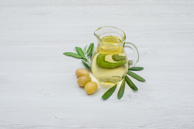 Green olives and olive oil in a glass jar with olive leaves side view on white wood plank