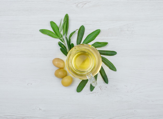 Free photo green olives and olive oil in a glass jar with olive tree branch top view on white wood plank
