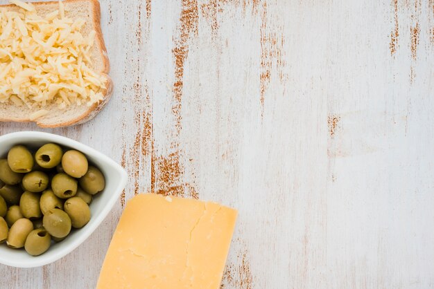 Зеленые оливки в белой миске; тертый сыр на хлебе над белым столом