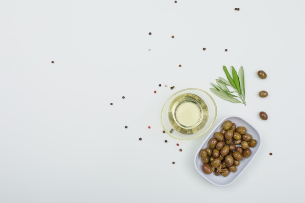 Green olives with a jar of olive oil, spices and olive leaves in a white plate on white, top view.