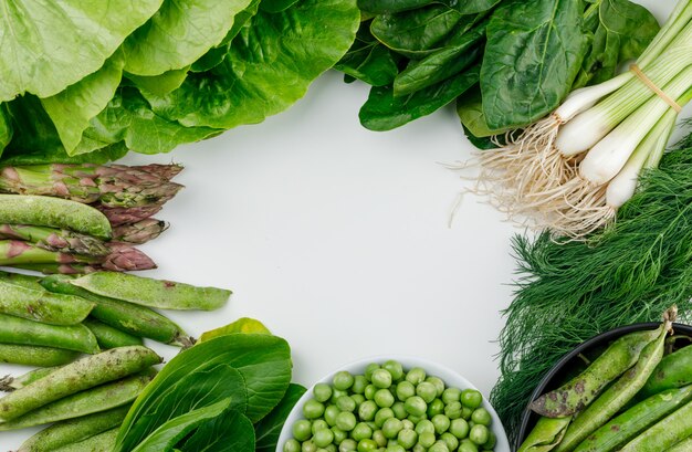 Green peas, pods in bowl and saucepan with spinach, sorrel, dill, lettuce, asparagus, green onions flat lay on a white wall