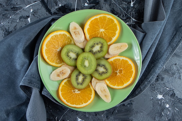 Зеленая тарелка нарезанного апельсина, киви и банана на мраморной поверхности.