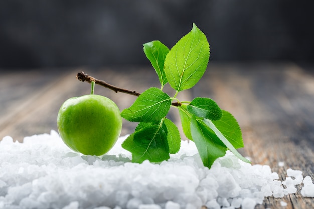 Green plum with branch and salt crystals on wooden and grungy wall, side view.