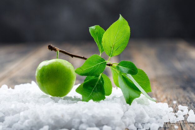 Green plum with branch and salt crystals on wooden and grungy wall, side view.