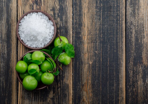 Green plums in a clay plate with salt in bowl and leaves top view on a wooden wall