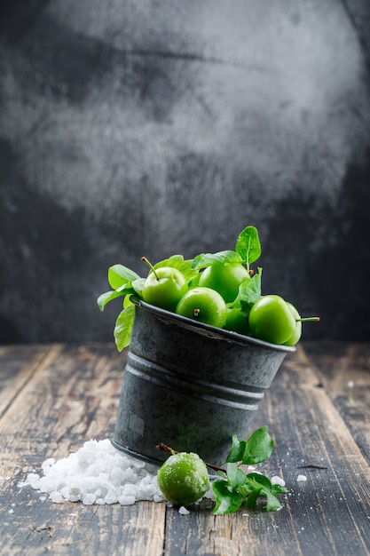 Green plums in a mini bucket with salt crystals, leaves side view on wooden and misty wall