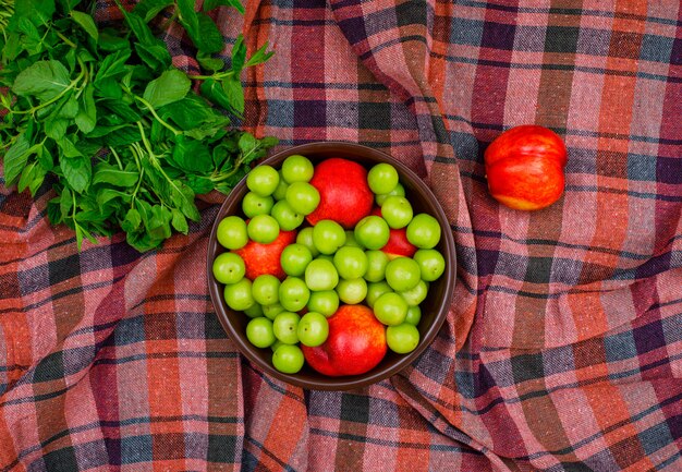 Green plums and peaches in a clay bowl with green leaves flat lay on picnic cloth