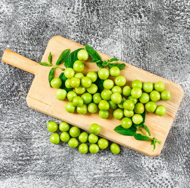 Green plums with leaves in a wood cutting board flat lay on grungy grey