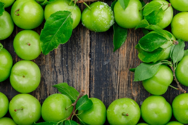 Green plums with leaves on wooden wall, flat lay.