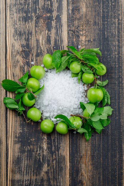 Green plums with salt crystals, leaves top view on a wooden wall