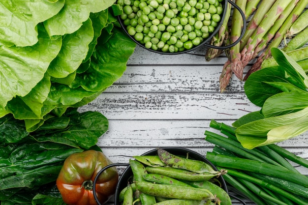 Green pods, peas in saucepans with asparagus, tomato, sorrel, spinach, lettuce, green onion top view on a wooden wall