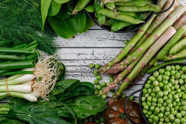 Green pods, peas with tomato, dill, sorrel, asparagus, green onion, bok choy in saucepans on wooden wall, top view.