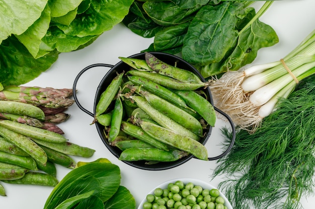 Green pods with peas, spinach, sorrel, dill, lettuce, asparagus, green onions in a saucepan on white wall, top view.