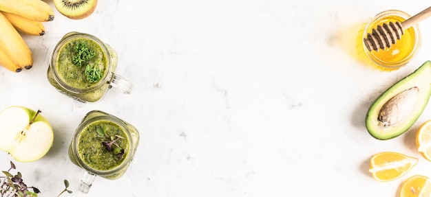 Green smoothie of vegetables and fruits with ingredients in glass jars on a white background, top view, copy space.