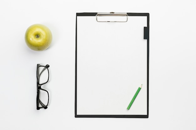 Free photo green whole apple with eyeglasses and green pencil on white clipboard over white office desk