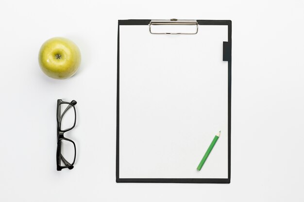 Green whole apple with eyeglasses and green pencil on white clipboard over white office desk