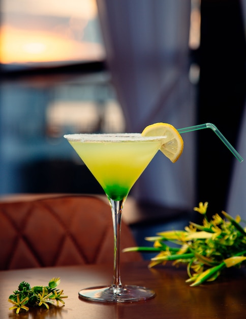 Green yellow cocktail with lemon slice and flowers around.