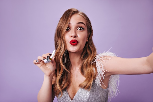 Greeneyed lady in festive dress holding party pipe and whistling Young woman in silver rop looks surprised and taking selfie on purple background