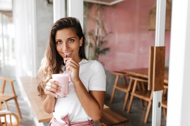 Зеленоглазая улыбающаяся женщина в белой футболке пьет молочный коктейль, сидя в кафе
