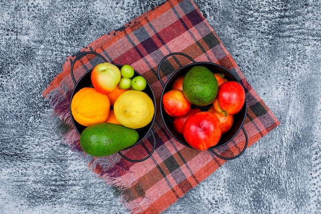 Greengages, pesche, avocado e agrumi in due casseruole nere su tela da picnic e grunge. disteso.