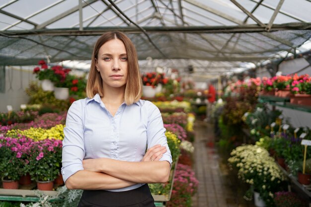 Greenhouse owner posing with folded arms having many flowers and a colleague holding a pot with pink chrysanthemums under glass roof