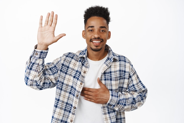 Greeting. Smiling friendly african american guy tell his name, raise hand and put arm on chest, introduce himself, say hello, making promise, standing on white.