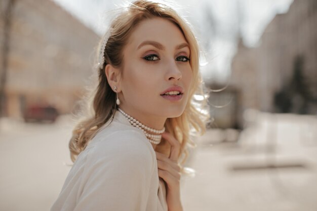 Grey-eyed beautiful woman with makeup looks into camera. Pretty girl in white blouse and pearl necklace poses outside