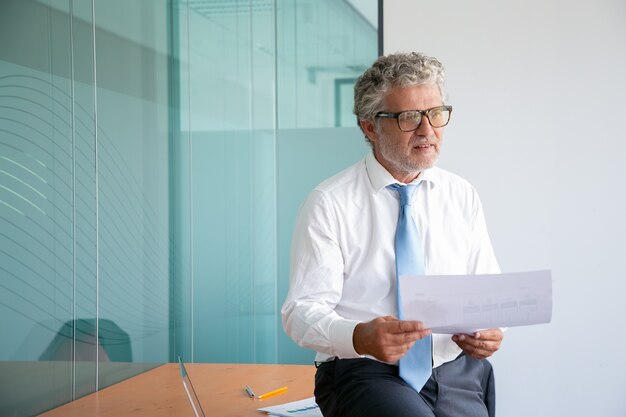 Grey-haired Caucasian businessman sitting on table and holding paper