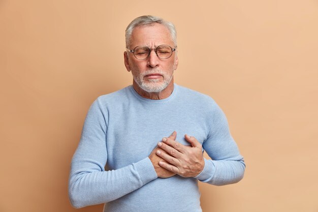 Grey haired displeased bearded old man has sudden painful spasm in chest closes eyes and presses hands to heart poses against beige wall