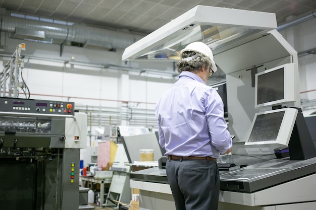 Седовласый инженер-заводчик в каске и очках стоит у промышленного оборудования, используя цифровое устройство