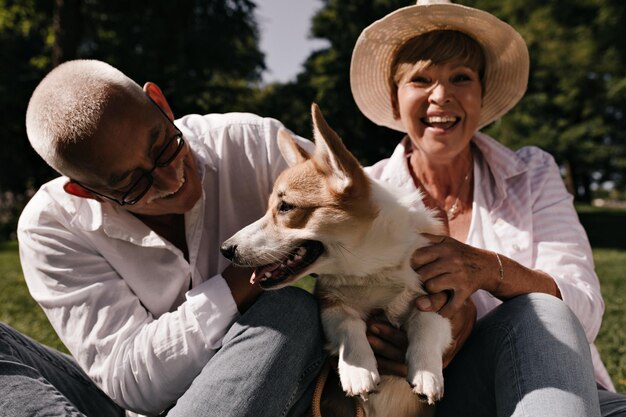 Grey haired man in eyeglasses and white shirt smiling and posing with cute corgi and blonde woman in hat and light blouse outdoor