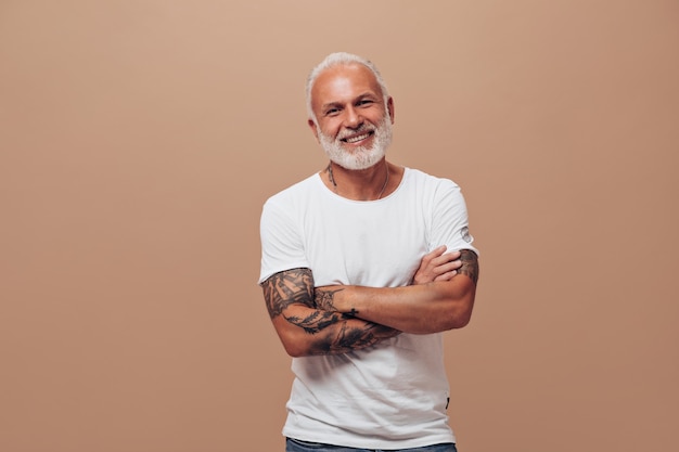 Grey haired man in white T-shirt poses on beige wall