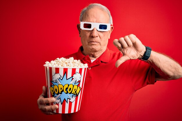 Grey haired senior man wearing 3d movie glasses and eating popcorn over red background with angry face negative sign showing dislike with thumbs down rejection concept