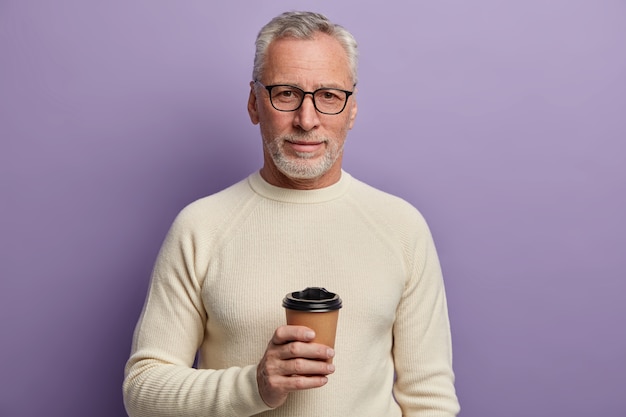 Free photo grey haired senior man wears transparent glasses and white sweater, stands and cools hot beverage, enjoys pleasant conversation, poses against purple background.