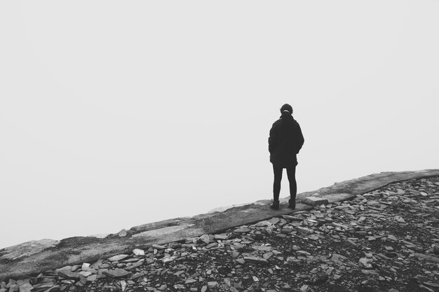 Grey scale shot of a person standing on the edge of a cliff looking in the white void