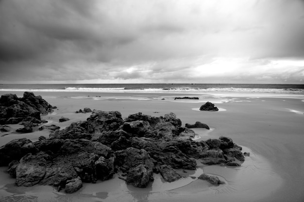 Greyscale of the beach surrounded by rocks and a wavy sea under a cloudy sky at daytime