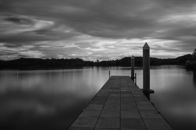 Greyscale of a pier on the sea surrounded by islands covered in greenery under a cloudy sky