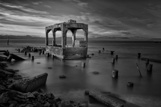 Greyscale shot of building ruins surrounded by wooden logs in the sea under the beautiful cloudy sky