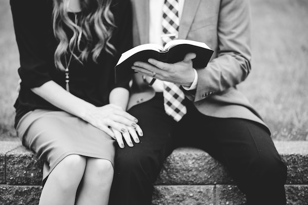 Greyscale shot of a couple wearing formal clothes while reading a book together in a garden