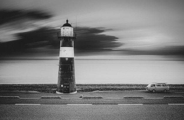Greyscale shot of a lighthouse on the road with a van on the side and the sea on the