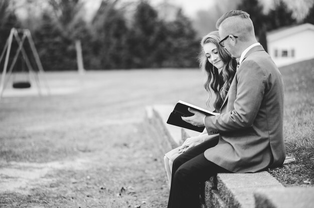 Greyscale shot of a male and a female wearing formal clothes while reading together in a garden