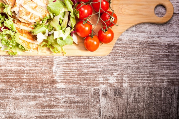 Petto di pollo alla griglia su insalata di taglio fresco accanto a un grappolo di pomodori su una tavola di legno